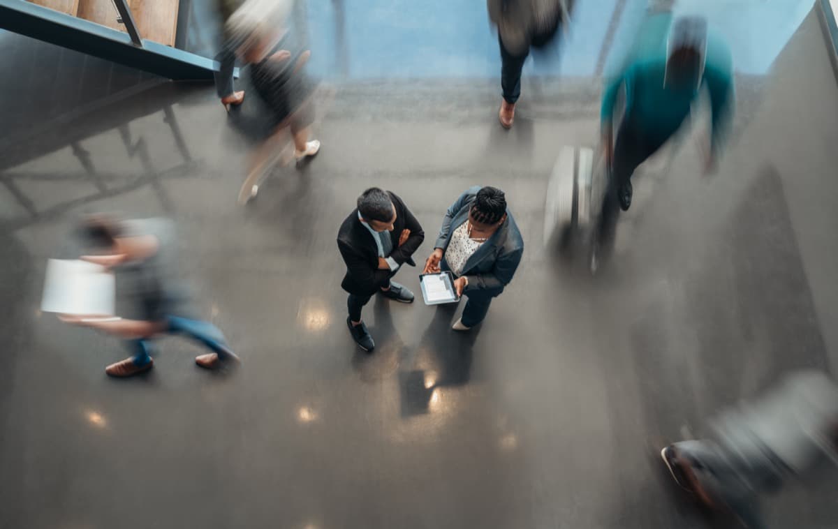 Two business people standing in the lobby of an office looking at a tablet while people are walking past in a blur; return to office concept