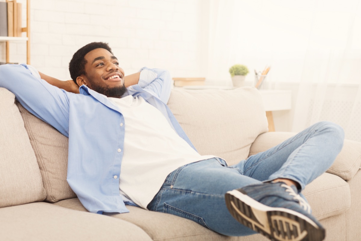 Young man relaxing at home, holding hands behind head and smiling on sofa; on-demand pay concept