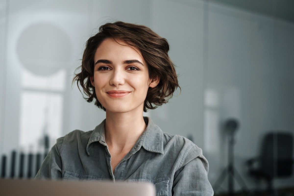 Image of young joyful woman smiling while working with laptop in office; employee development concept